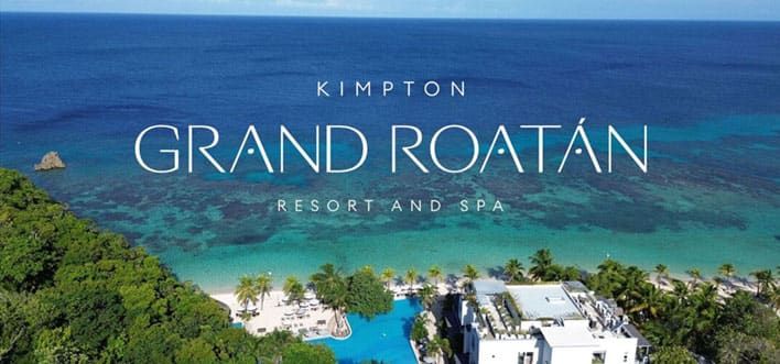 Aerial view of Kimpton Grand Roatán Resort and Spa with white buildings, palm trees, pool, and clear blue ocean in the background.