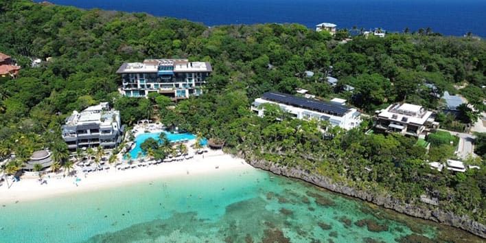Aerial view of beachfront resorts surrounded by lush greenery, with clear turquoise water and white sand on a tropical coastline.