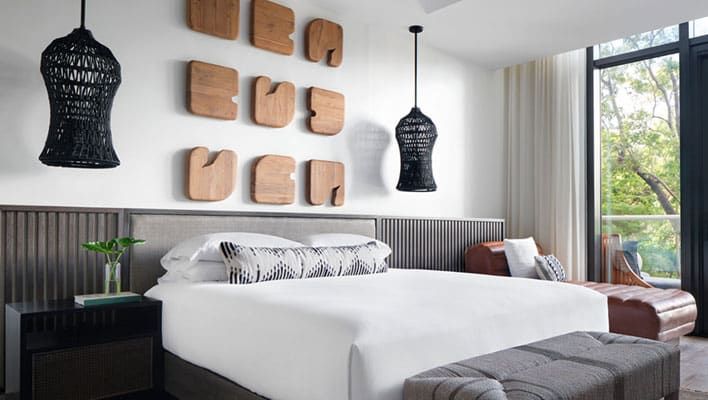 Kimpton Grand Roatán room with a white bed, geometric wooden wall art, two black pendant lamps, large windows, a brown chaise lounge, and a gray upholstered bench at the foot of the bed.