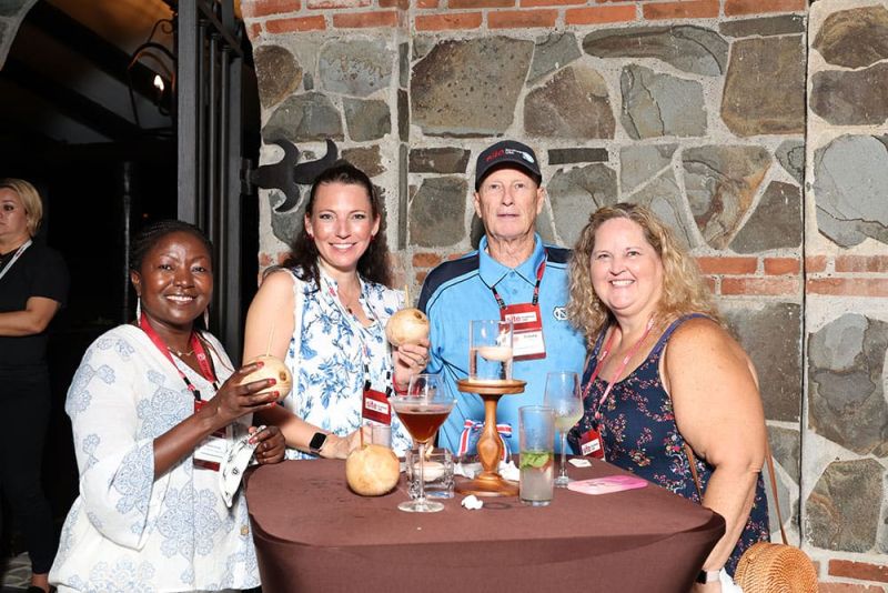 Four people standing around a round table with drinks, posing for a photo in front of a brick and stone wall.