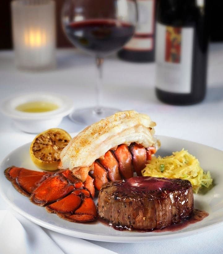 A plated meal featuring a grilled lobster tail and a steak topped with sauce beside sliced lemon and a side dish, served with a glass of red wine.