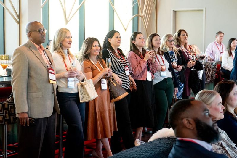 A group of people with name tags stand and sit, attentively facing forward during SITE Southeast’s 2025 Kickoff event. Glasses and drinks are visible on a table nearby.