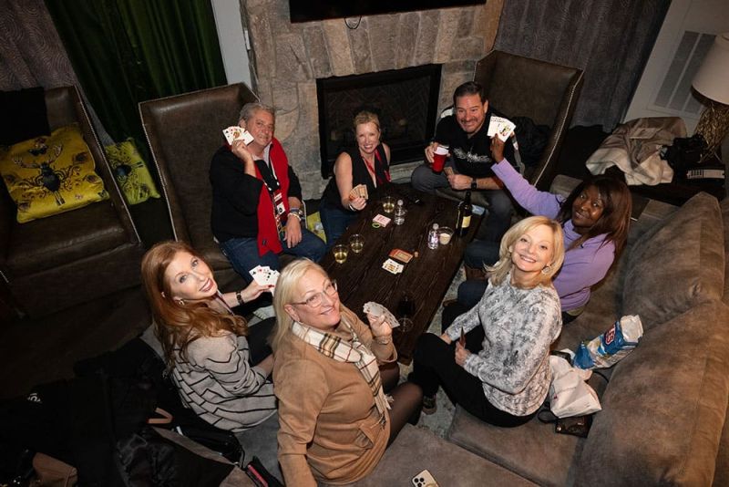 Seven people are sitting around a table playing cards, smiling and holding their hands of cards up. Drinks and snacks are on the table.