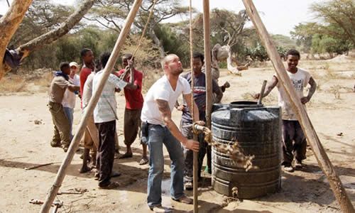 A group of people, including Doc Hendley in a white t-shirt operating a pulley, are working together to draw water from a well as part of the Wine to Water initiative in a dry, rural area. Trees and shrubs are visible in the background.