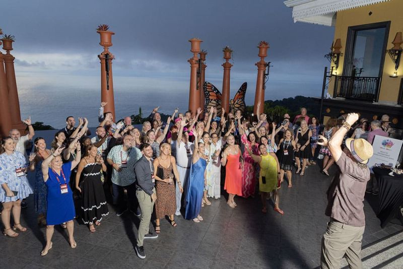 A group of people posing outdoors on a terrace, some raising their arms. The background shows the ocean and a cloudy sky.