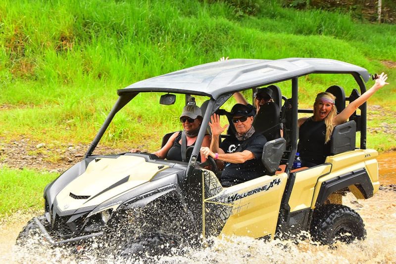 Four people in an off-road vehicle driving through water, with one woman waving and smiling.