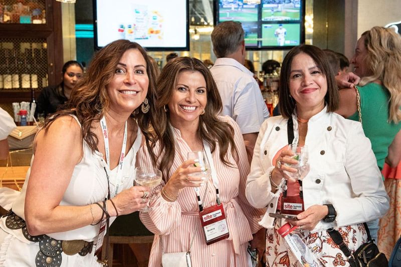 Three women with name badges stand together, smiling and holding drinks at SITE Southeast’s 2024 Southern Supper. Screens displaying sports can be seen in the background.