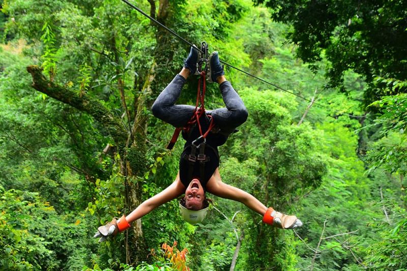 Person ziplining upside down with arms outstretched, surrounded by lush green forest.